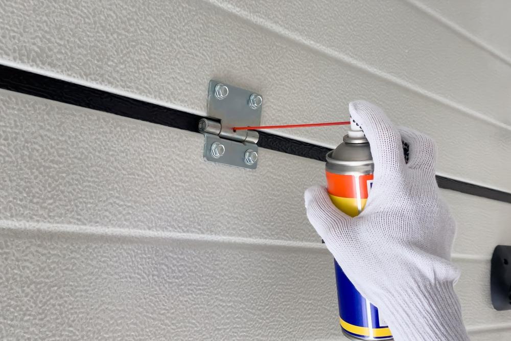 A white-gloved hand using a spray bottle to lubricate garage door hinges.
